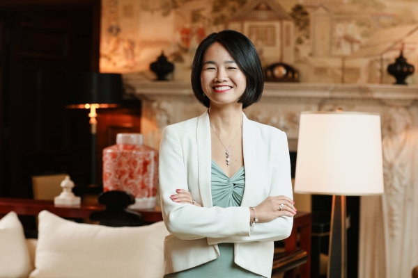 A portrait of Sally Maier-Yip smiling while stood in front of aa fireplace, sofa and lamps. Sally is an Asian woman wearing a whie jacket and green dress stands with folded arms.